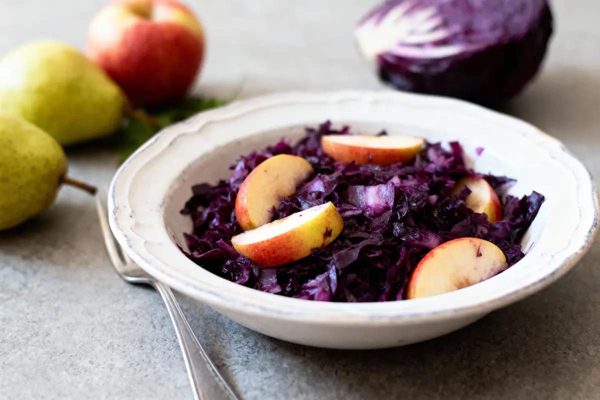Salade de chou rouge et pommes dans un bol blanc, avec poires et chou en arrière-plan.