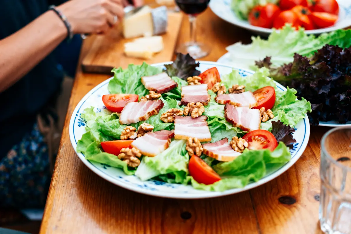 Salade de laitue, tomates, noix, lardons. Assiette sur table en bois avec fromage et vin en arrière-plan.