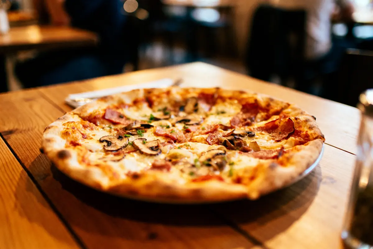 Pizza garnie champignons et jambon sur table en bois, ambiance de restaurant en arrière-plan flou.