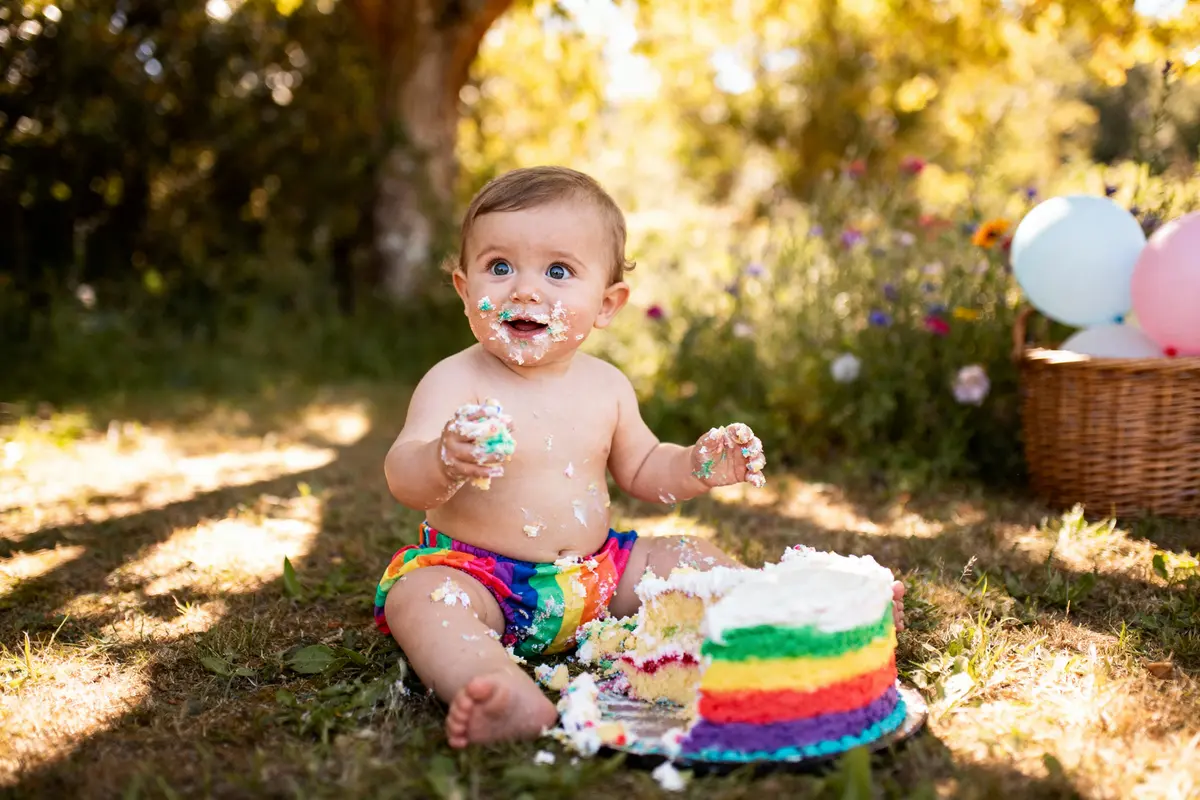 Bébé souriant, couvert de gâteau coloré, assis sur l'herbe près d'un panier et de ballons.