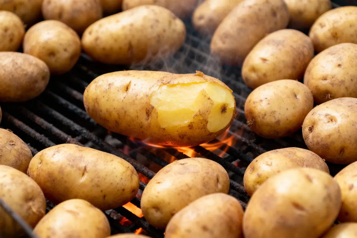 Pommes de terre grillées sur barbecue, avec peau légèrement pelée, flammes visibles en dessous.