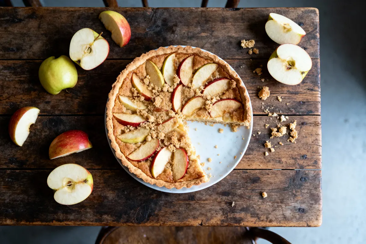 Tarte aux pommes avec tranches sur table en bois, entourée de morceaux de pommes.