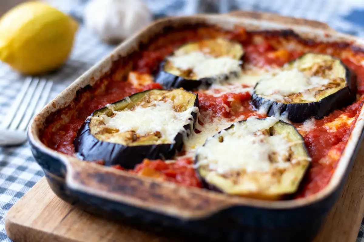Plat gratiné avec tranches d'aubergine, sauce tomate, fromage fondu, sur nappe à carreaux. Citron et ail visibles.