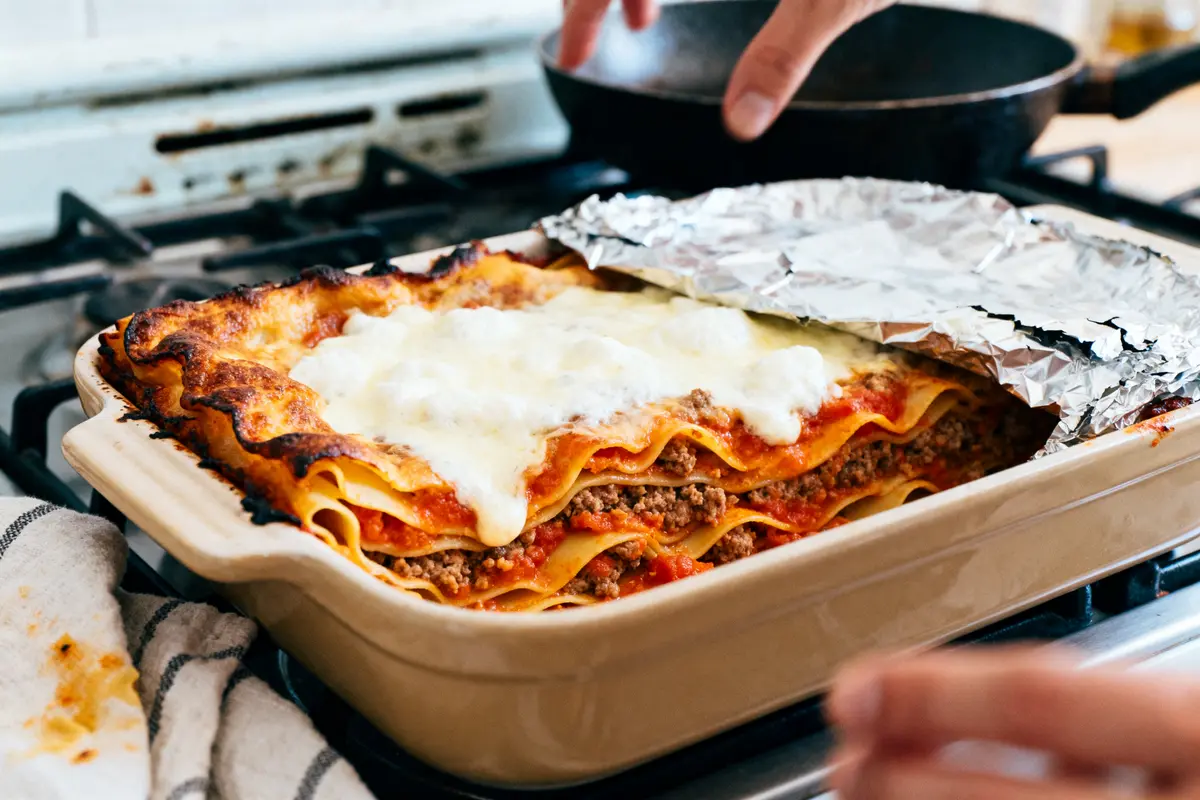 Lasagne maison au fromage gratiné dans un plat, main retirant du papier aluminium.