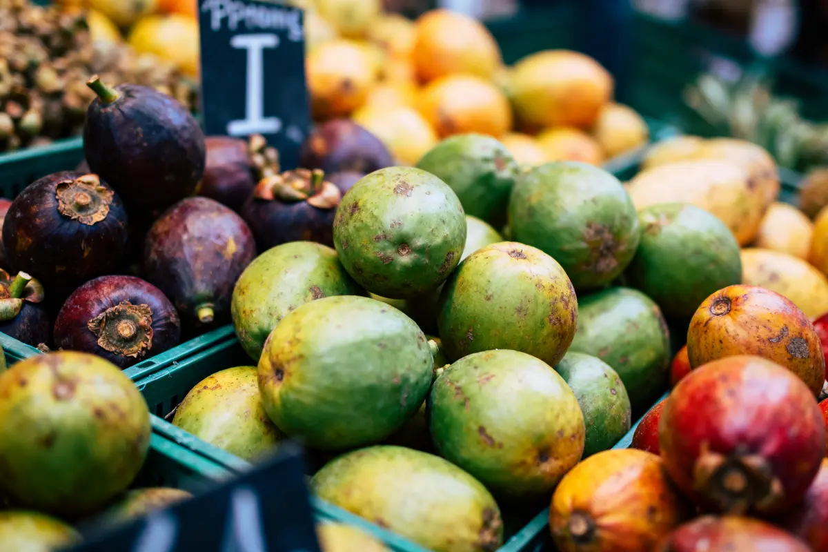 Fruits variés au marché : mangoustans, mangues et autres, en bacs verts, avec étiquette de prix visible.